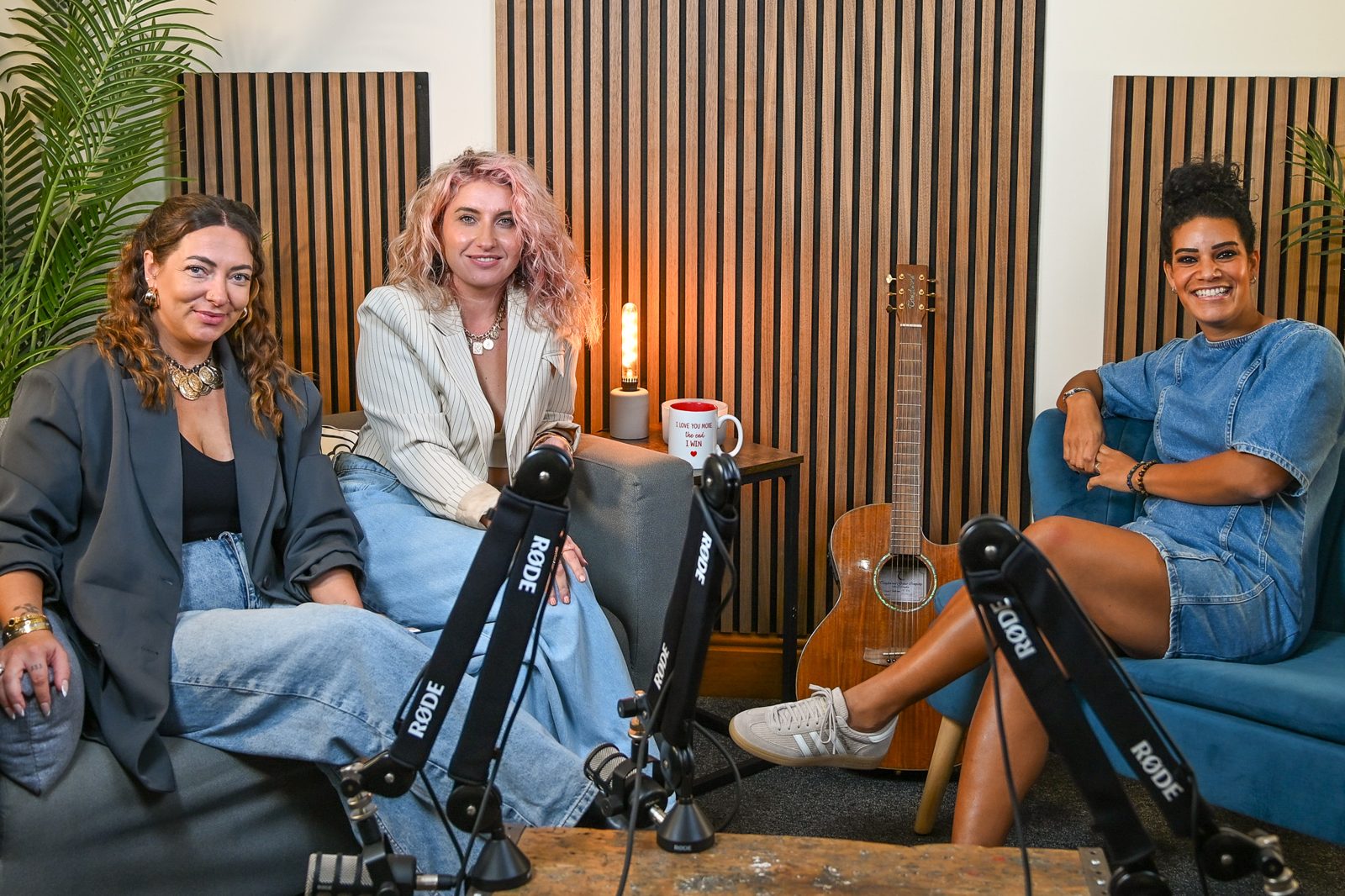 Three podcast hosts smiling on the sofas of the wood-panel set, with microphones in front of them